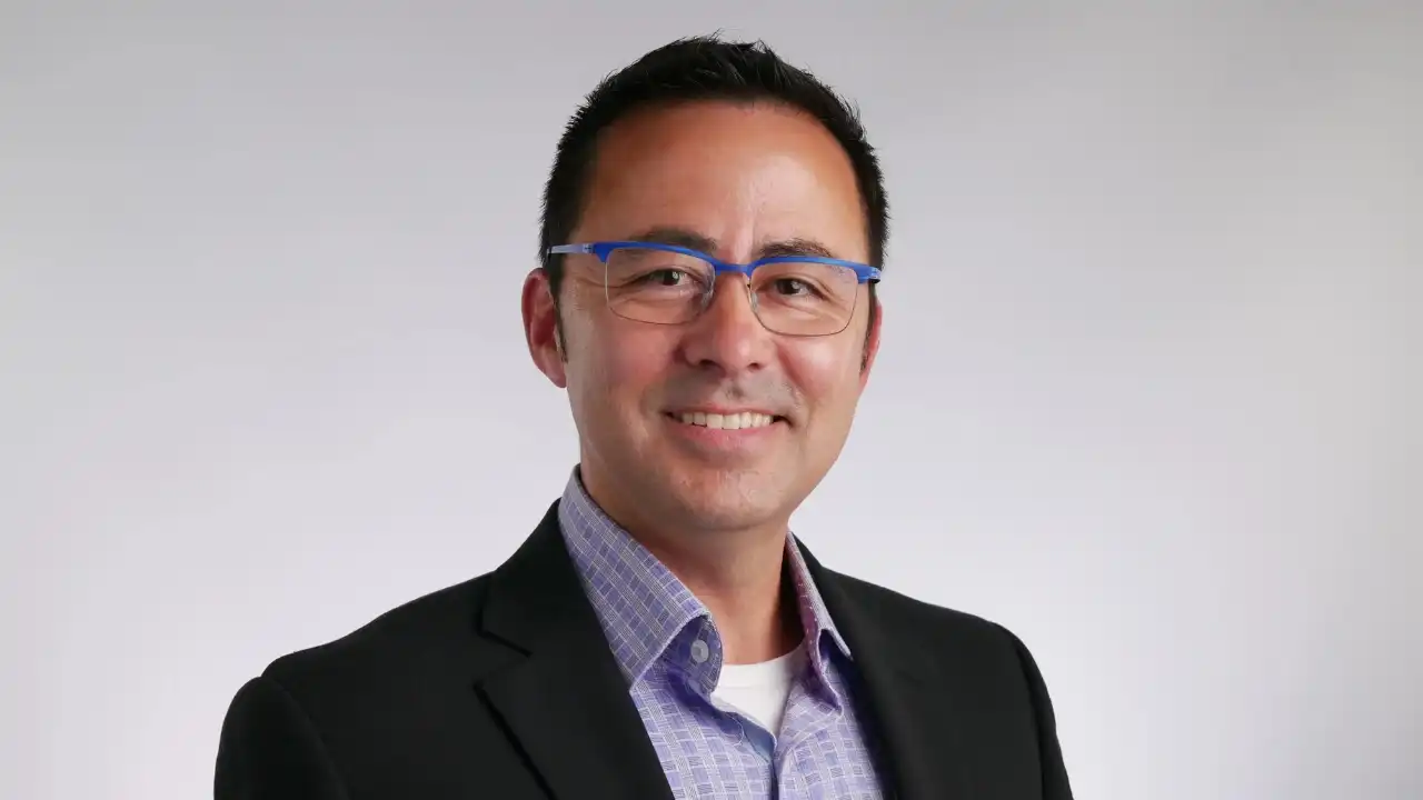 Portrait photo of a smiling man wearing blue-rimmed glasses and a black blazer.