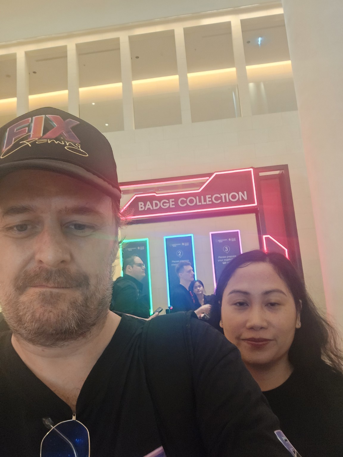 Two Fix Gaming Channel attendees taking a selfie beside the illuminated badge-collection area.