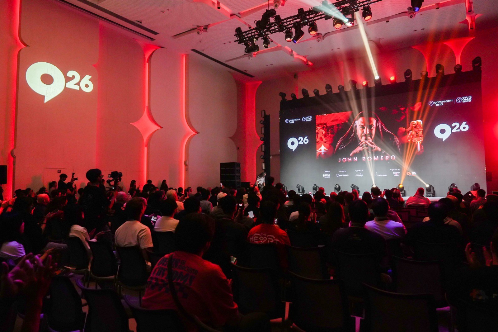 An audience seated in a red-lit auditorium beneath a stage screen displaying John Romero's name at gamescom asia x Thailand Game Show 2026.