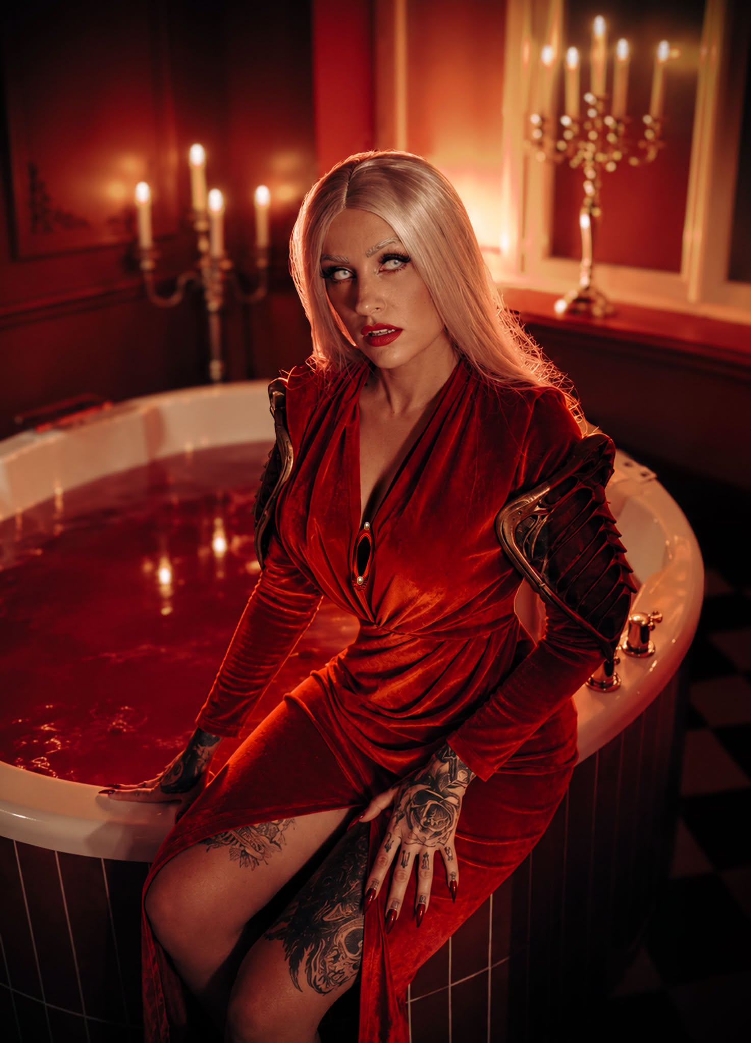 Kax in a red dress and dark shoulder armor beside a candlelit bathtub filled with red liquid.